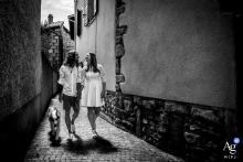 Brindas, Rhône — The newlyweds walk hand in hand with their dog down an alleyway, sharing a kiss as the sun sets behind them, casting long shadows in this black-and-white, urban wedding portrait.