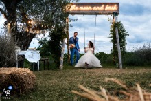 Rimini — Outdoors, the bride sits playfully on a swing while the groom stands behind her, creating a lighthearted and joyful wedding portrait in the natural setting.