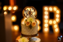   At Mariënhof Amersfoort, a beautifully decorated wedding cake stands in front of large, illuminated “MR & MRS” letters, with fresh flowers adorning the top tier and adding to the festive atmosphere.