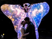 At Ca' Borghese in Ameglia, La Spezia, Italy, the bride and groom share an intimate embrace beneath a luminous winged performer, whose glowing costume creates a dramatic guardian-like backdrop that transforms the portrait into a striking blend of romance 