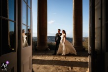 At Prior Park in Bath, England, the bride and groom stand between elegant columns at sunset as their reflection is photographed through open paned-glass doors, creating a layered portrait rich with symmetry and warm evening light.