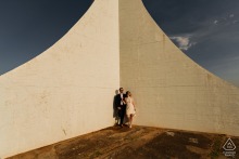 A couple from Brasilia poses cheerfully in the bright sunlight, standing at the corner of an artistic backdrop. 