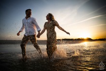 A couple joyfully runs through shallow water at Crane Beach, Ipswich, MA, during golden hour, creating romantic reflections.