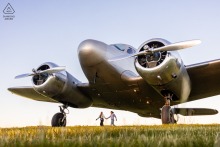 Engaged couple holds hands beneath vintage airplane wings at Cape Cod Airfield, symbolizing timeless romance.