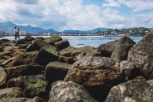 SAINT-JEAN-DE-LUZ, France — A couple stands on the rocks overlooking the bay of Saint-Jean-de-Luz, with the city and the La Rhune mountain in the distance for their engagement portrait.