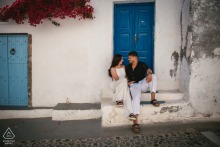 SANTORINI, Greece — A couple relaxes on a stoop of a classic white building with a vibrant blue door. This engagement portrait captures their quiet connection amidst the iconic streets of Santorini.
