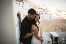 SANTORINI, Greece — A couple embraces above a village, with the groom-to-be hugging his fiancée from behind. This romantic engagement portrait captures their affection in Greece.