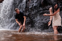 An engaged couple playfully splashes in a Duluth, MN waterfall, surrounded by rocks and cascading water.
