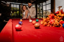 Engaged couple plays pool at Vale dos Ventos, Rio dos Cedros, SC, smiling at each other, bouquet and balls visible.