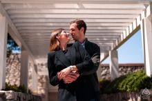 Por — A couple hugs as he wraps her up from behind at a sunlit, white-painted wood trellis, creating a bright and intimate portrait.