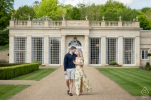 A couple walks together in front of the orangery at Rushton Hall, symmetrically framed by green lawns on either side. The elegant architecture creates a refined backdrop for their engagement portrait.