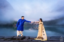 A couple stands on stone pavers at a Lake Louise lookout, reaching their hands toward each other. Soft fog surrounds them, creating a dreamy and romantic atmosphere for their engagement portrait.