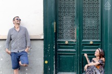 In Paris, France, a couple enjoys a casual, relaxed session on a Parisian street. She sits on the stoop of a green painted doorway while he stands nearby, capturing the charm of everyday romance.