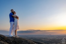 At Les Rochers du Carnaval, a couple perches on rocky outcrops at a scenic lookout. He embraces her from behind, both gazing out over the landscape in this sunset engagement portrait.