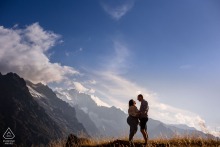 Minimalist portrait at Lac du Pontet, France, showing an engaged couple positioned at the bottom of the frame beneath vast blue sky, wispy clouds, and towering mountains.