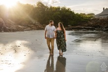 The warm glow of golden hour at Wingaersheek Beach frames a couple walking hand-in-hand. The shallow water creates a mirror effect on the sand beneath their feet, enhancing the scene.