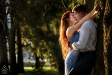 In Maciel, Santa Fe, a couple poses beside a park tree as she kisses him, both bathed in warm, orange sunlight for their portrait.