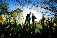 In Philadelphia, a couple leans in through tall grass and flowers, about to kiss, with trees and a blue sky above, captured in a portrait that highlights the photographer’s mastery of location lighting.