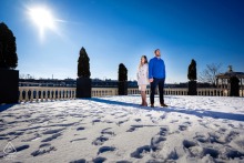 At Fairmount Waterworks in Philadelphia, a couple stands on snow-covered ground with footprints, captured against a blue sky and backlit by the sun.