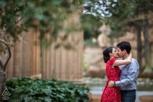 An engaged couple kisses in a garden setting surrounded by trees and wooden fences during a portrait session in San Francisco, California.