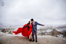 An engaged couple hugs with one arm and stretches out their other arms while kissing atop a scenic overlook during a pre-wedding photo session in Cappadocia, Göreme, Nevşehir, Turkey.