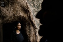 A man stands silhouetted in the foreground while his fiancée, softly illuminated, stands behind him framed by a cave doorway during a pre-wedding photo session in Cappadocia, Göreme, Nevşehir, Turkey.