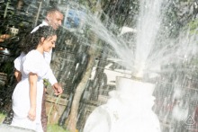 Couple walking together in Porto Alegre, their affectionate connection glimpsed through the distorted water spray of a fountain that partially obscures them.