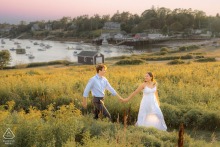 Couple walks hand in hand through a coastal meadow in Harpswell, Maine, surrounded by yellow flowers and the sea, bathed in warm sunlight during their romantic engagement session.