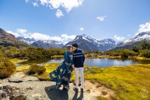Couple face each other on Key Summit, Routeburn Track, New Zealand, both wearing knit caps against the mountain lake’s biting wind, sunlight illuminating their profiles with nature’s grandeur behind them.