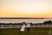 A couple walks hand in hand along the shoreline near Nobska Lighthouse in Falmouth, MA at sunset, surrounded by soft pastel skies and calm light, capturing timeless Cape Cod romance.
