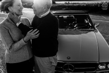 An elderly couple stands close beside a classic car at Dowsers Beach, Osterville, MA, sharing a tender glance in a timeless black and white portrait that celebrates enduring love and genuine connection.