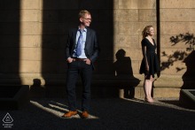 Engagement portrait of a couple standing in golden sunlight, their shadows stretching across the large stone walls of an old world building in San Francisco.