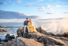 An engaged couple perches on rocky coastal ledge as tide crashes against the shoreline in Kennebunkport, Maine.