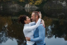 An engaged couple stands closely aligned along the riverbank while watching the water flow past in Metz, Moselle, France.