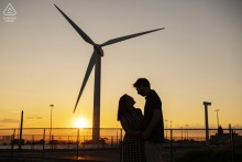 An engaged couple stands silhouetted against a colorful sunset sky near a towering wind turbine on Deer Island in Boston, Massachusetts.