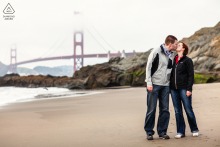 Engaged couple kisses on a San Francisco beach with the Golden Gate Bridge visible beyond the rocky shoreline during their portrait session in California.