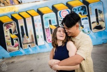 he couple hugs and laughs together in front of the famous Chicago mural in Logan Square, Chicago, Illinois, celebrating their engagement with a vibrant and local piece of street art.