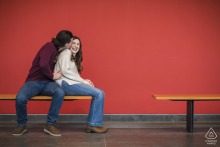 Inside the Cleveland Museum of Art Atrium in Cleveland, Ohio, the engaged couple playfully kisses while sitting on a bench, a vibrant red background bringing warmth and energy to their affectionate pose.