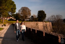Walking side by side along the historic castle walls in Gorizia, Italy, a couple enjoys quiet conversation while posing for an engagement portrait with sweeping views and historic stone architecture.