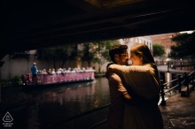An engaged couple embraces beneath a River Walk bridge during a pre-wedding portrait session in San Antonio, Texas, as a river barge passes behind them.