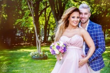 Pre-wedding engagement portrait of a couple embracing in a sunlit garden in Bucharest, Romania, with the bride holding a bouquet of colorful flowers.