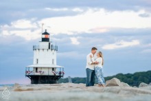 Pre-wedding engagement portrait of a couple embracing on a scenic breakwater with a lighthouse in South Portland, Maine.