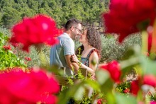 A couple kisses in a lush garden framed by bright red blooms during their engagement session in Jaén, Andalucía, Spain.