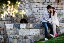An engaged couple sits on a stone wall in Napa Valley, California, pausing for a kiss during their outdoor engagement photo session