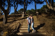A couple stands together on a wooded park staircase during a pre-wedding engagement session in San Francisco, California, surrounded by towering trees and natural beauty.