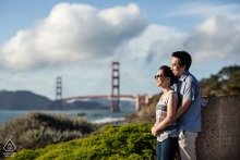 A couple enjoys a pre-wedding engagement portrait overlooking San Francisco Bay, with the Golden Gate Bridge and bright white clouds creating a timeless California backdrop.
