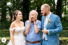 On an outdoor wedding day in Echteld, the Netherlands, the bride and groom stand beside an emotional older man who covers his face, reflecting a poignant family moment filled with love.