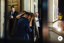 Viewed through a window at Two Temple Place, London, the couple shares a kiss in the rain, sheltering under the groom’s coat and creating a romantic scene despite the weather.