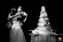 Bride and groom surrounded by wedding guests in a group hug while cutting cake at Akra Barut Otel, Antalya, Turkey, with a towering cake at right.