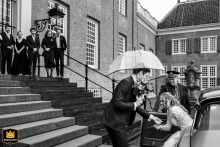 At Slot Zeist, the groom, holding an umbrella, helps his bride from the car, while smiling guests await at the top of the stairs in this black and white shot.